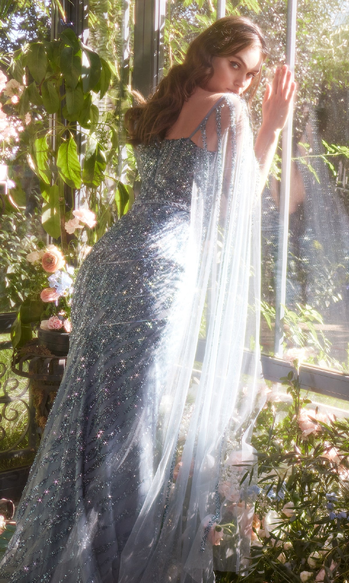 A woman wearing the Andrea & Leo A1370 long formal dress stands by a sunlit window surrounded by lush green plants and pink flowers.